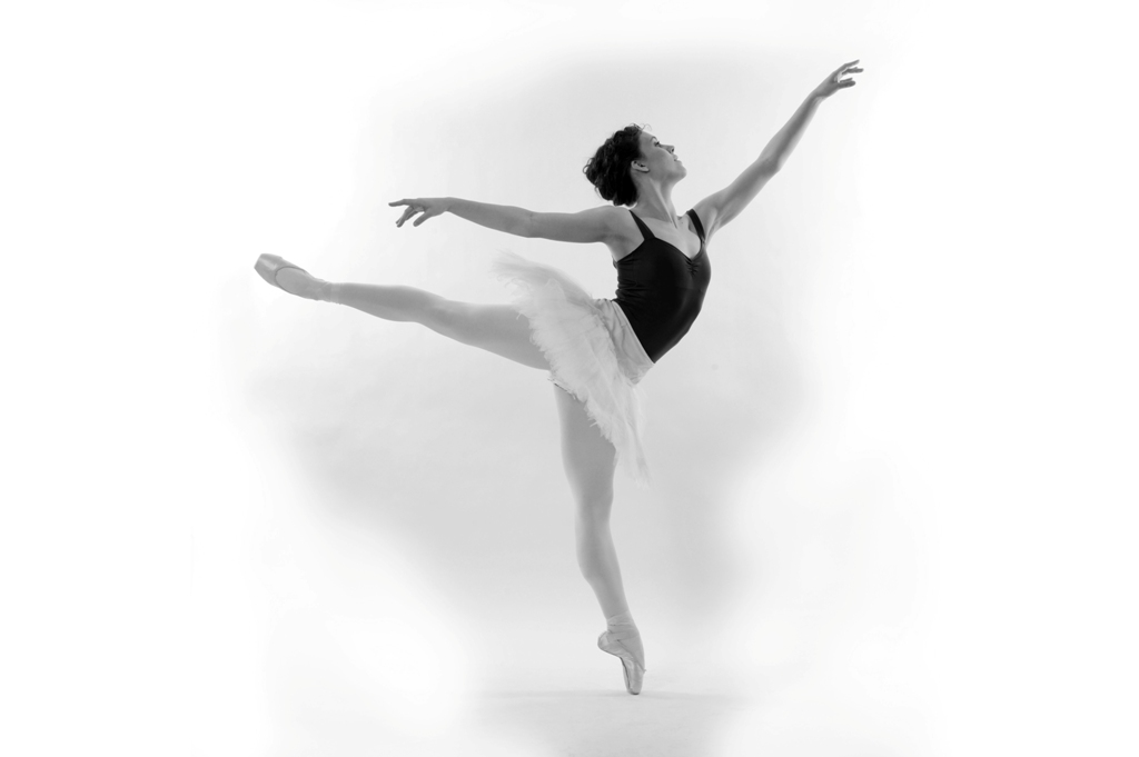Black and white photo of Natasha in arabesque, wearing a dark leotard, white tutu, and pointe shoes